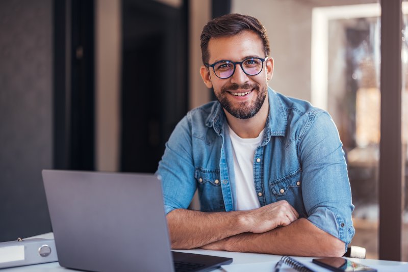 A smiling and bearded man wearing glasses and sitting in front of a laptop